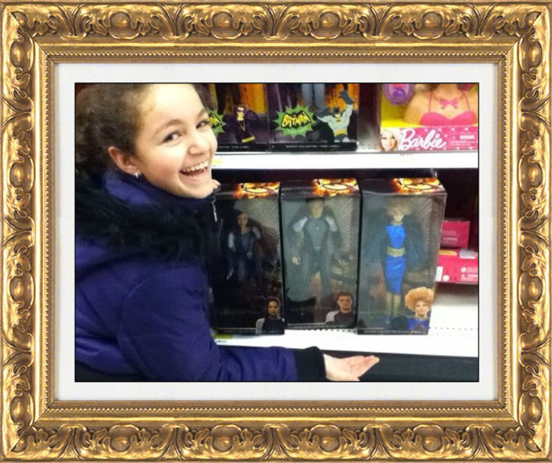 Image is of author Julia Bifulco, younger, smiling in front of a shelf of The Hunger Games dolls in a store.