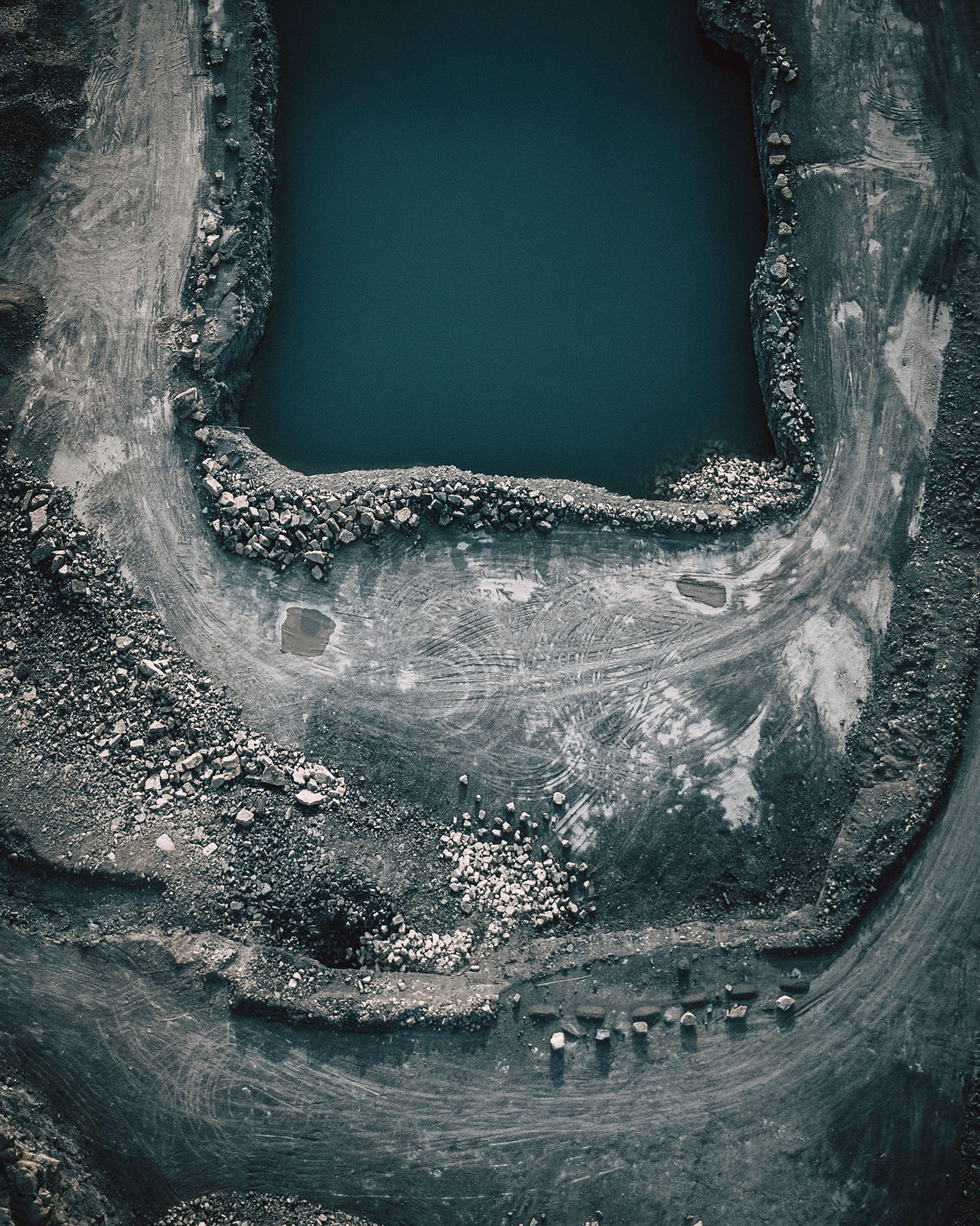 Image is an aerial view photograph of a small body of blue water surrounded by grey rocks.